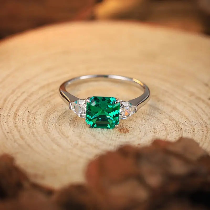 Silver ring with a square-cut emerald gemstone flanked by two smaller diamonds.