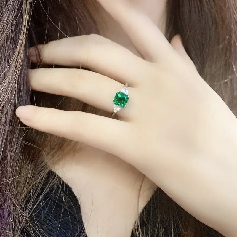 Emerald ring with a square-cut green gemstone and diamond accents on a pale hand.