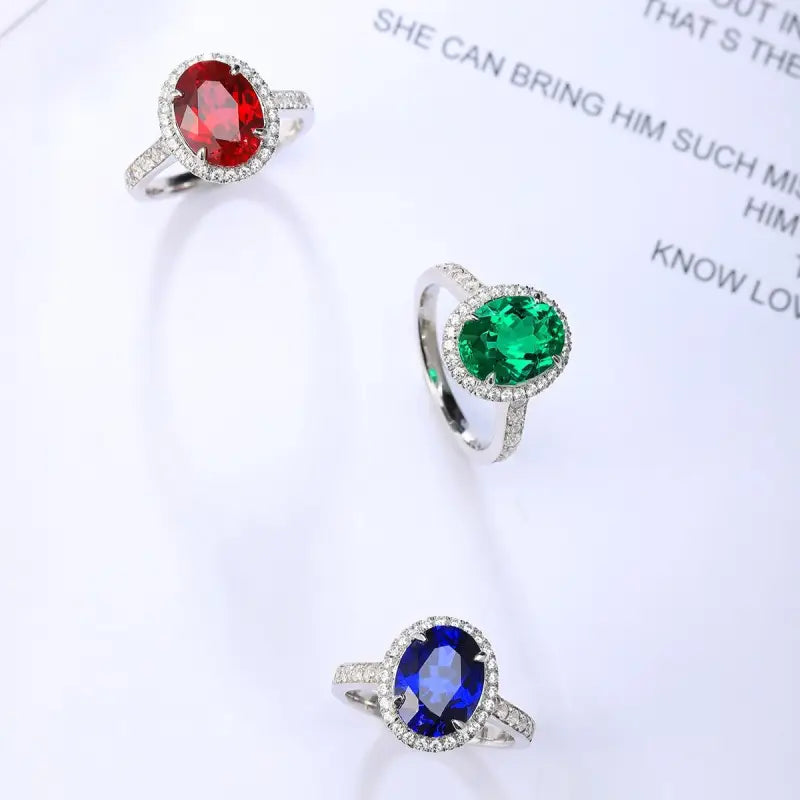 Three gemstone rings featuring red, green, and blue oval-cut stones surrounded by diamond halos.