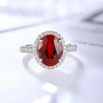 Load image into Gallery viewer, Oval-cut ruby ring with diamond halo and band set in white metal.
