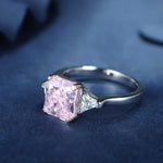 Load image into Gallery viewer, Engagement ring featuring a square-cut pink diamond center stone flanked by two smaller triangular diamonds on a silver band.
