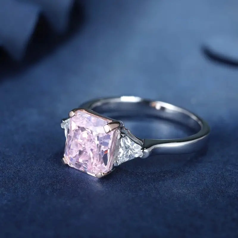 Engagement ring featuring a square-cut pink diamond center stone flanked by two smaller triangular diamonds on a silver band.