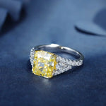 Load image into Gallery viewer, Engagement ring featuring a square-cut yellow diamond center stone flanked by smaller white diamonds on a split shank.
