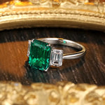 Load image into Gallery viewer, Emerald-cut green gemstone ring with diamond side stones set in a white metal band.
