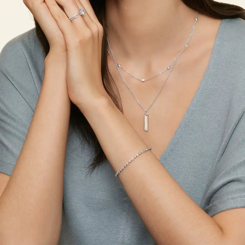 Delicate silver jewelry set including necklaces, bracelet, and ring worn by a person in a gray top.