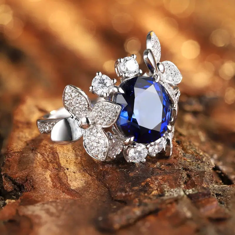 Ornate silver ring featuring a deep blue gemstone surrounded by diamond-encrusted floral designs.