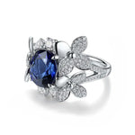 Load image into Gallery viewer, Ornate silver ring featuring a deep blue gemstone surrounded by diamond-encrusted flower-shaped designs.
